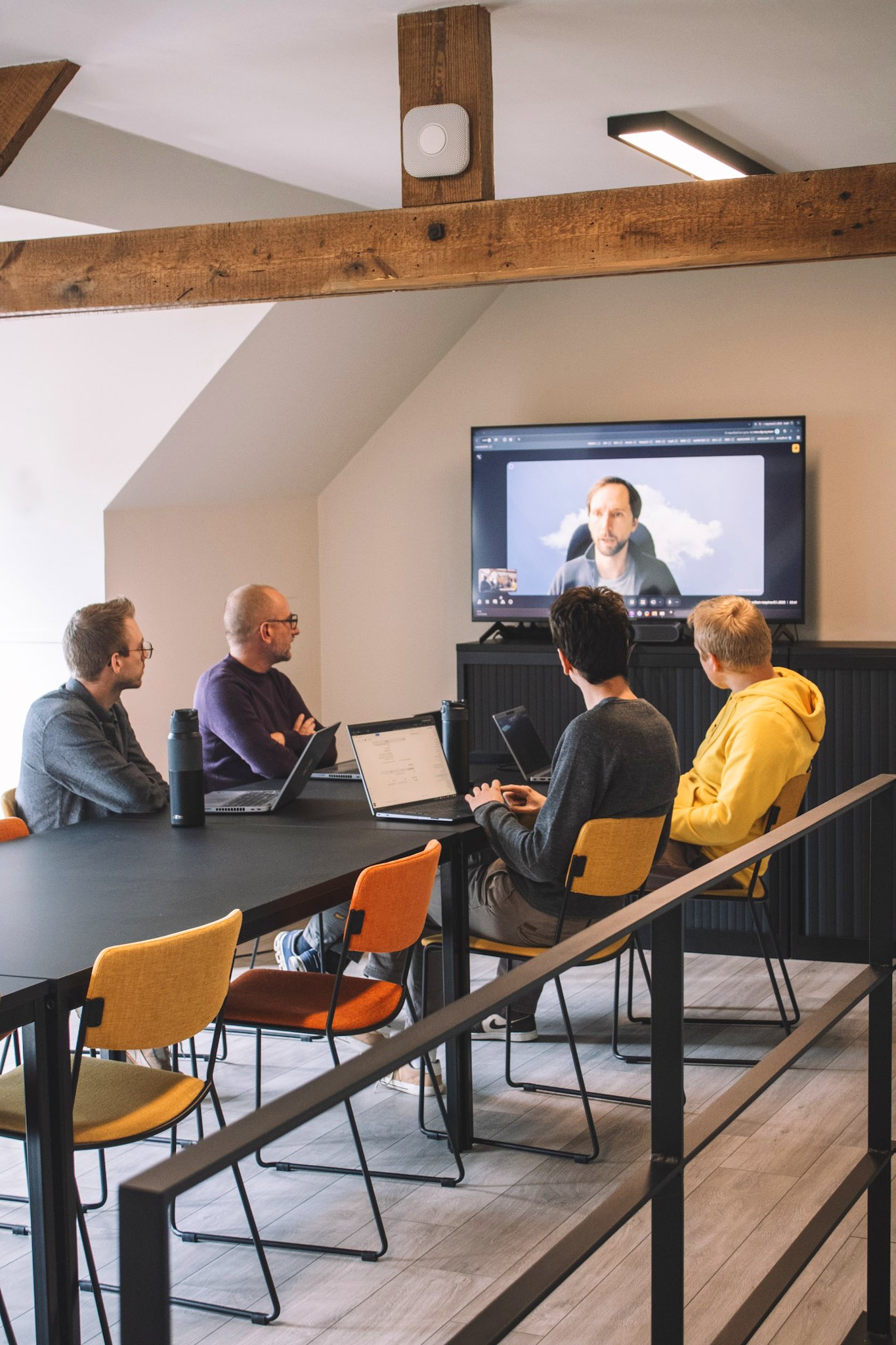 Mad IT team in overleg tijdens hybride vergadering met collega op scherm, werkend aan digitale IT-oplossingen in moderne vergaderruimte met laptops en samenwerkingstools.