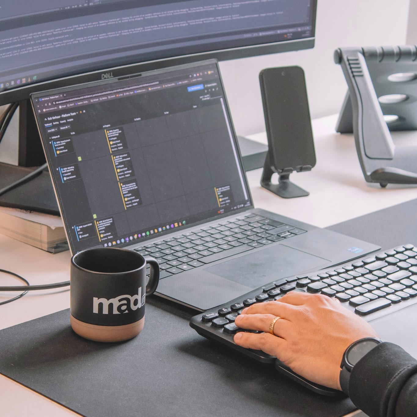 Hand van Mad IT-medewerker op toetsenbord naast laptop met programmeercode op scherm en Mad IT-koffietas - moderne werkplek voor .NET softwareontwikkeling.