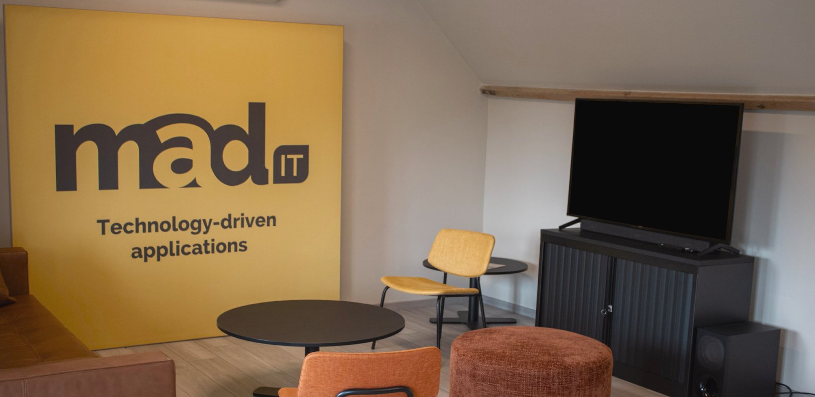 Moderne kantoorruimte van Mad IT met gele wand met logo en slogan "Technology-driven applications", zithoek met designstoelen, ronde tafels en flatscreen-tv op zwart tv-meubel - creatieve werkplek voor technologie en softwareontwikkeling.